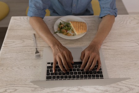Mid section of young Asian male executive using laptop while eating lunch in modern officeの写真素材