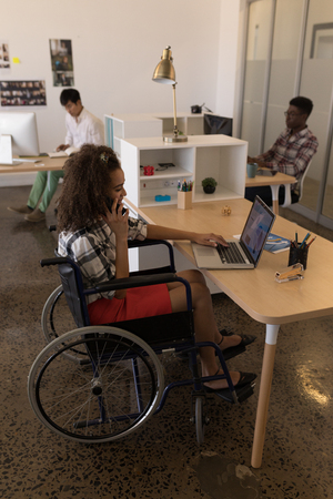 Side view of young disabled mixed-race female executive using laptop while talking on mobile phone at desk in modern office.の写真素材