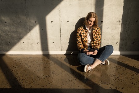 Front view of beautiful young Caucasian female executive using mobile phone sitting on the floor in modern officeの写真素材