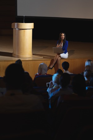 High view of beautiful businesswoman sitting at the side of the stage and having laptop in her hand in front of audienceの写真素材