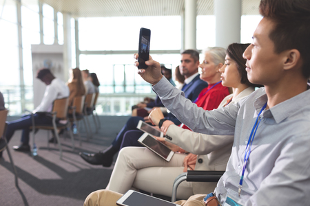 Side view of young Asian businessman clicking photo with mobile phone in a business seminar in office buildingの写真素材