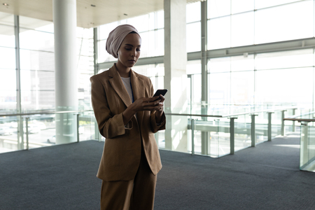 Front view of mixed-race businesswoman using mobile phone in office lobby against view on street in backgroundの写真素材