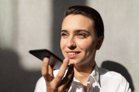 Portrait of pretty young Caucasian businesswoman talking on mobile phone standing in modern office. She is smilingの写真素材
