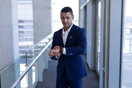 Front view of handsome young mixed-race businessman using smartwatch standing in modern officeの写真素材