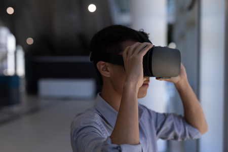 Side view of young Asian male executive using virtual reality headset standing in modern officeの写真素材