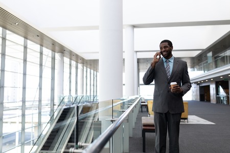 Front view of young African-American businessman with coffee cup talking on mobile phone standing in modern office. He is smilingの写真素材