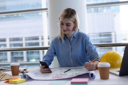 Front view of happy blonde young Caucasian female architect using mobile phone at desk in modern...