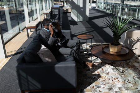 Side view of young African-American businessman sitting on the sofa and using virtual reality headset in modern officeの写真素材