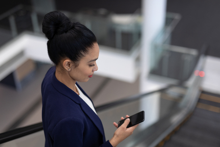 Side view of pretty young mixed-race businesswoman using mobile phone on escalator in modern officeの写真素材