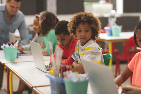 Side view of school kids using laptop in classroom while teacher teaching in backgroundの写真素材