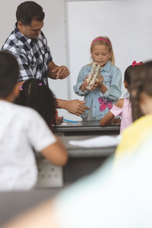 Front view of a Caucasian schoolgirl holding a skull in her hands and showing this one to others school kids sitting in front of her and his teacher next to her behind a deskの写真素材