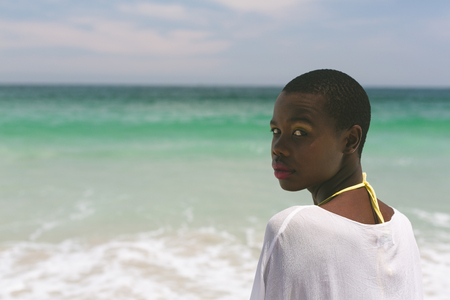 Rear view of African-american woman standing on beach on a sunny day while looking at cameraの写真素材