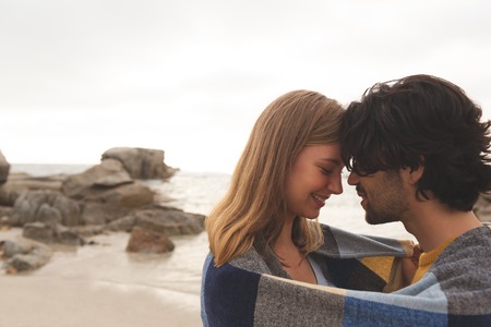 Side view of young Caucasian couple wrapped in blanket kissing each other standing at beachの写真素材