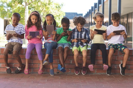 Front view of multi ethnic students using digital tablet while sitting on brick wall at corridor in schoolの写真素材