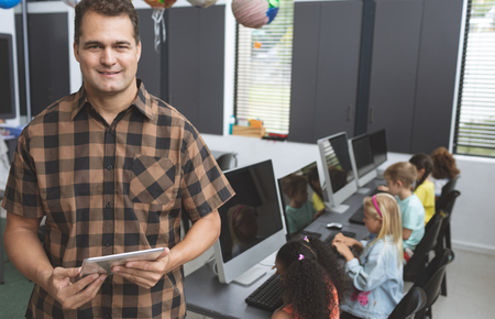 Portrait of happy schoolteacher holding digital tablet in classroom at school with school kids working in a computer room in backgroundの写真素材