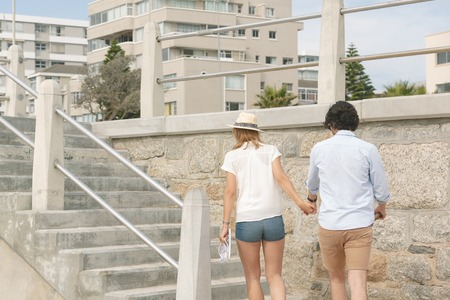 Rear view of young Caucasian couple walking hand in hand near sea side at promenade on a sunny day