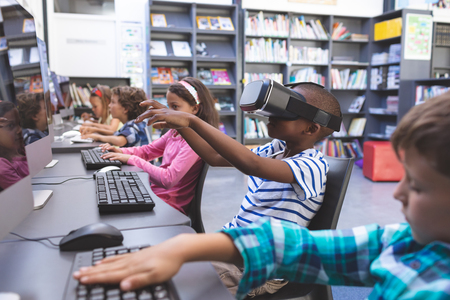 Side view of African-american schoolboy using virtual reality headset while his schoolmaes working...