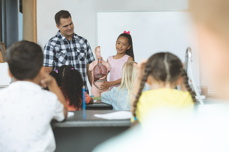 Front view of a schoolgirl explaining to his classmates the functioning of body human thanks to a dummy skeleton in classroom at school with his teacher next to herの写真素材