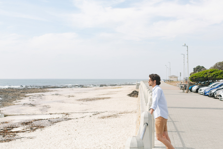 Side view of young Caucasian man standing near sea side at promenade on a sunny day. He are looking awayの写真素材