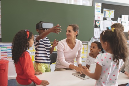 Front view of schoolboy using virtual reality headset at school in classroom with his classmates and her teacher next to himの写真素材