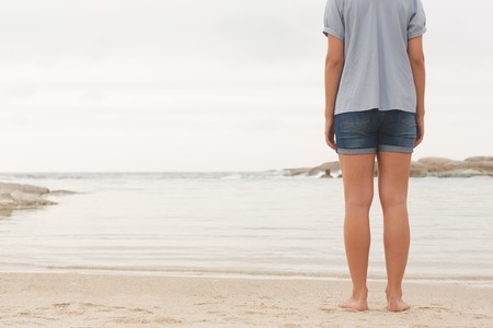 Low section of young Caucasian woman standing at beach on the sand. She is looking the sea