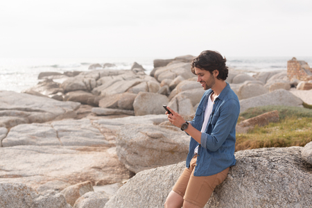 Side view of young Caucasian man using mobile phone at beach while leaning on rock. He seems happyの写真素材