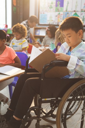 Side view of schoolboy studying in classroom while sitting at wheelchair in classroom at schoolの写真素材