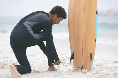 Side view of young mixed-race male surfer tying surfboard leash on his leg at beachの写真素材