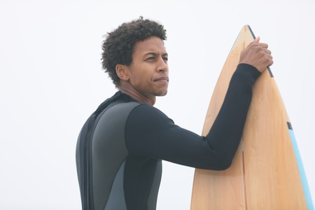 Side view of thoughtful young mixed-race male surfer holding surfboard on the beach. He is looking awayの写真素材