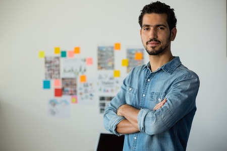 Portrait of confident attractive young mixed race man looking and smiling at camera with arms crossed standing in officeの写真素材