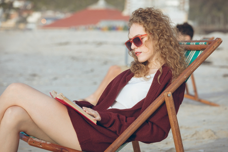 Front view of beautiful young Caucasian woman with sunglasses reading book while sitting on sun lounger at beach on sunny day.の写真素材