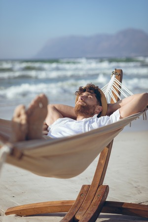 Front view of handsome young Caucasian man relaxing on hammock at beach on a sunny dayの写真素材