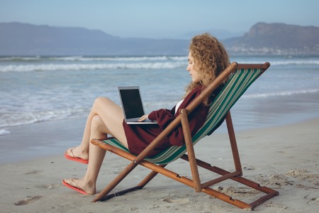 Side view of beautiful young Caucasian woman using laptop while sitting on sun lounger at beachの写真素材
