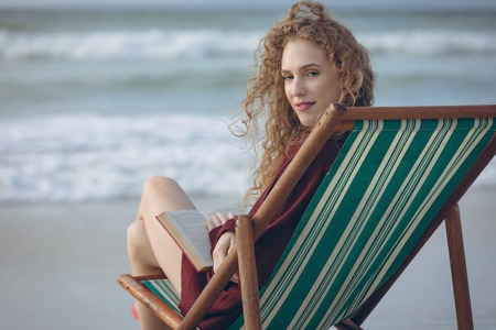 Portrait of beautiful young Caucasian woman holding book while sitting on sun lounger at beach. She...