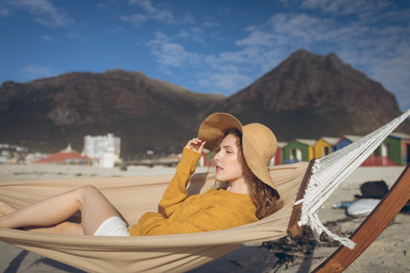 Side view of beautiful young Caucasian woman lying on hammock at beach on sunny day. She wears a hatの写真素材
