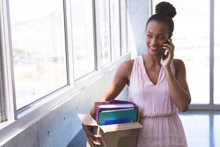 Front view of young mixed-race businesswoman talking on mobile phone while holding stuffs in the modern officeの写真素材