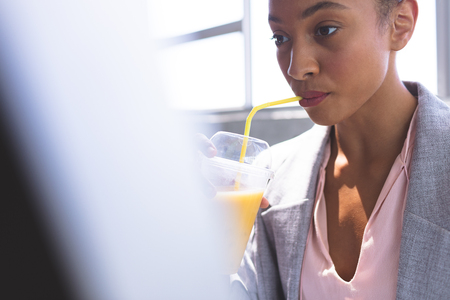 Front view of young mixed-race businesswoman having milkshake while working on computer at desk in the officeの写真素材