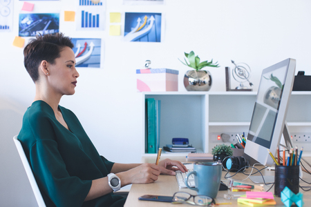 Side view of beautiful Mixed-race female executive writing on notepad while working on computer at desk in the officeの写真素材