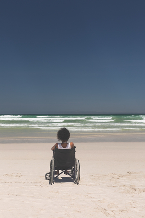 Rear view of young mixed-race woman disable sitting on wheelchair at beach on a sunny day. She is looking awayの写真素材