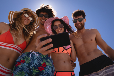 Low angle view of group of Multi ethnic friends taking selfie with mobile phone at beach in the sunshine against blue skyの写真素材