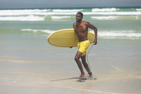 Front view of handsome fit African American male surfer with a surfboard running out of the sea water on the beach on a sunny dayの写真素材