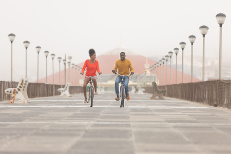 Front view of happy Multi-ethnic couple riding bicycle at promenade on a sunny dayの写真素材