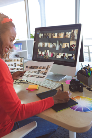 Side view of attentive African American female graphic designer working at desk while holding paper with photographs  in a modern office. She is smiling.の写真素材