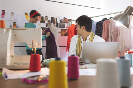 Front view of Asian male fashion designer talking with Mixed-race female designer while working on laptop in design studioの写真素材