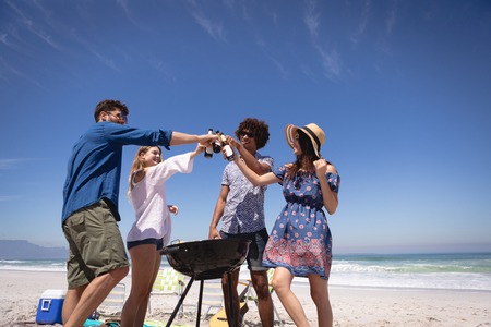 Low angle view of group of Multi ethnic friends toasting beer bottle at beach in the sunshine surrounding of barbecue against ocean waves in backgroundの写真素材
