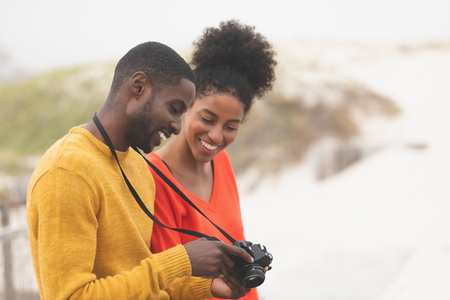 Side view of cute Multi-ethnic couple using digital camera while leaning against bench at dune. They are smilingの写真素材