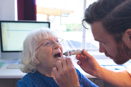 Side view of male Caucasian doctor checking senior female patient mouth with otoscope in clinicの写真素材
