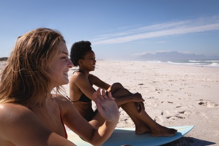 Side view of happy young multi ethnic women sitting on surfboards in front of the sea at beach in...