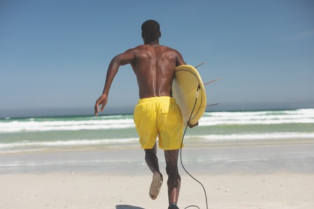 Rear view of African American male surfer with a surfboard running towards the sea water on a beach
