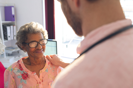 Rear view of a Caucasian confident male doctor interacting with female senior patient in clinicの写真素材